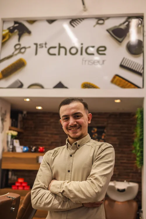 Ein junger Mann mit dunkelbraunen, nach vorne gekämmten Haaren steht mit einem Jeanshemd vor einer Ziegelmauer.
