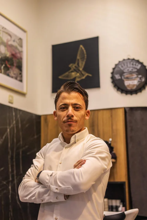 Ein junger Mann mit dunkelbraunen, nach hinten gekämmten Haaren steht mit einem weißen Hemd vor einer Ziegelmauer.
