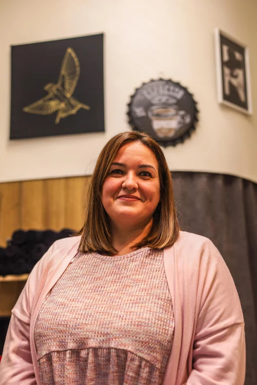 Eine junge Frau mit langen braunen, gewellten Haaren steht lächelnd in einem schwarzen Oberteil vor einer Ziegelmauer.
