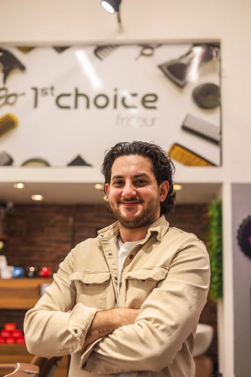 Ein Mann mit schwarzen zurückgegelten Haaren und einem Schnauz- und Kinnbart steht mit einem offenen, dunkelgrauen Hemd vor einer Ziegelmauer.
